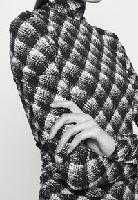 Czarnobiała tkanina w kratkę, wysoki kołnierz; długie rękawy z marszczeniem; fokus na wyciągniętej dłoni.