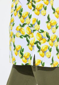 Tessuto bianco con un motivo di limoni e foglie verdi. Il design è vivace, mostrando limoni interi e a metà. Include un dettaglio con spacco laterale.