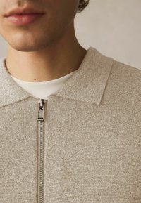 Beige Strickjacke mit strukturierter Oberfläche, mit einem Reißverschluss, einem weichen Kragen und einem weißen Hemd darunter.