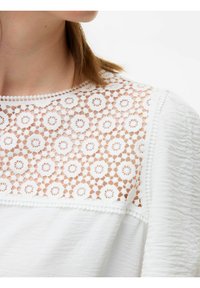 Vit blus med ett blommigt spetskrage med intrikata utskärningar och texturerad tyg. Dekorativ kant längs kanterna ger detaljer.
