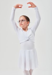 Weißer Langarmbody mit einer Drehung an der Taille, kombiniert mit einem fließenden, transparenten Rock. Das Model trägt eine Brille und hat die Haare zu einem Dutt gebunden.