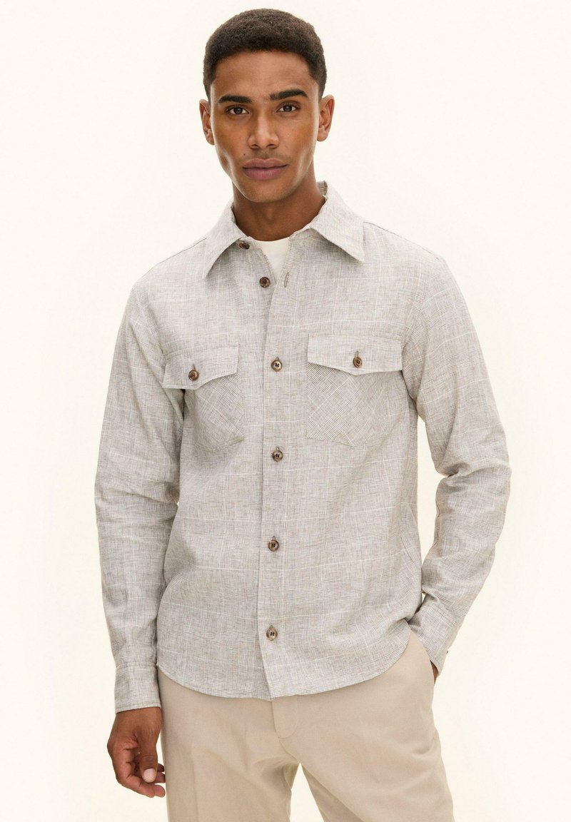 Homme portant une chemise beige texturée à boutons avec poches poitrine et un pantalon beige, debout avec une main dans la poche devant un fond uni.