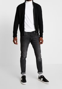 Man som bär en vit polotröja, svart texturerad zip-up jacka, blekta svarta jeans med uppvikta ben, vita strumpor och svarta sneakers med vita sulor.