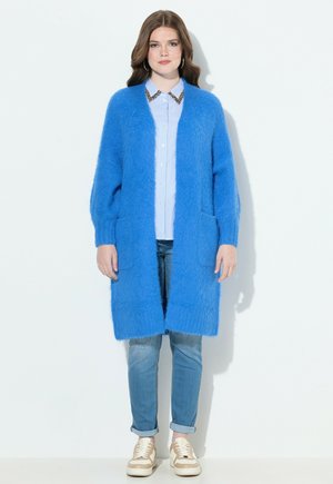 Cardigan azzurro brillante e fluffoso con apertura frontale, due tasche, indossato sopra una camicia a righe azzurra chiara, abbinato a jeans chiari e sneakers beige.