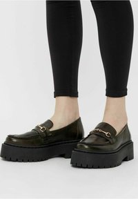 Donkergroene leren loafers met een stevige zwarte plateauzool en gouden kettingaccent. Gladde textuur met stikseldetails.