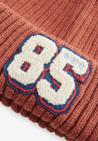 Stickad brun mössa med ribbad struktur, prydd med ett märke med siffran "85" i vit och blå ull på en blå bakgrund.