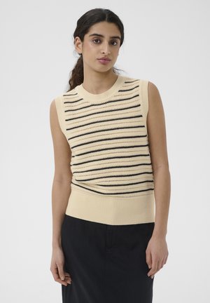 Jonge vrouw met donker haar die een mouwloze crèmekleurige geribbelde top met horizontale zwarte strepen en een zwarte rok draagt, staand tegen een witte achtergrond.