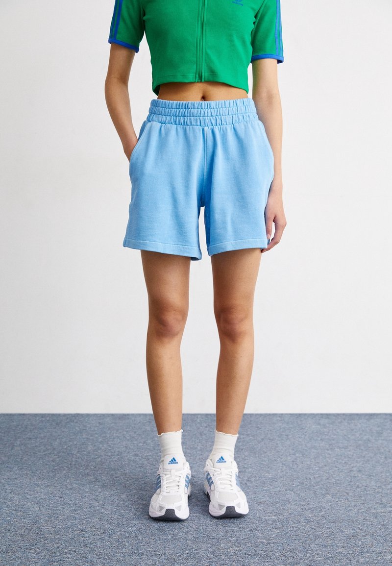 Light blue shorts with an elastic waistband and side pockets, paired with a green cropped top and white sneakers with blue accents.