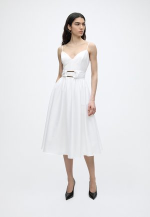 Woman with long dark hair wearing a white sleeveless midi dress with belt detail, standing and looking to the side, wearing black heels.