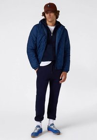Chaqueta navy acolchada sobre una sudadera oscura con capucha, combinada con pantalones de chándal oscuros y zapatillas deportivas azules. Incluye una gorra marrón y calcetines blancos.