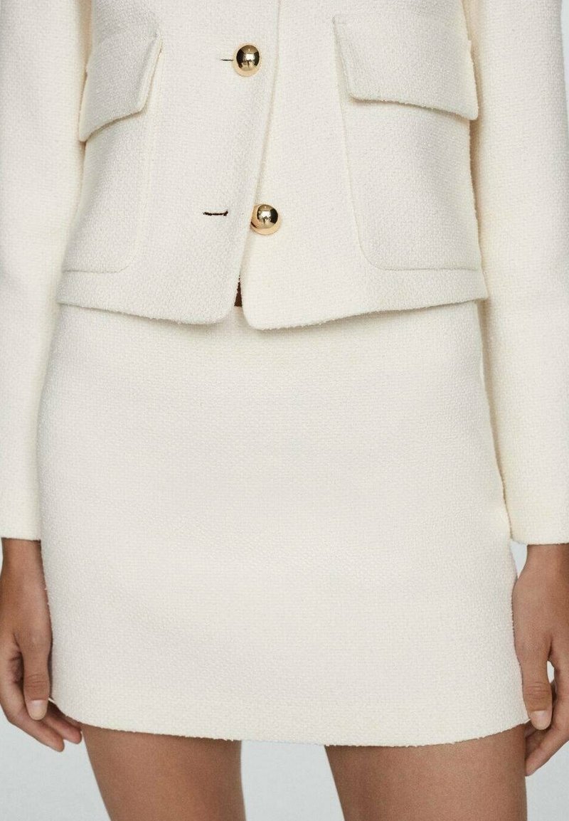 Blazer blanc texturé avec des boutons dorés et des poches avant, assorti à une mini-jupe blanche assortie. Les deux ont une finition lisse.