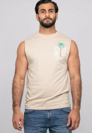 Homme aux cheveux foncés et à la barbe portant un débardeur beige avec le texte "Palm Beach" et un motif de palmier, associé à un jean bleu.
