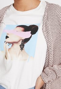 Vitt t-shirt med en grafisk bild av en kvinna med en blå bakgrund, rosa band över ögonen och svarta örhängen. Tillsammans med en texturerad beige kofta.