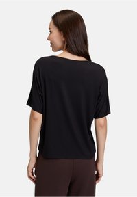 Czarny krótki top z luźnym krojem, szerokim dekoltem i gładką fakturą, zestawiony z ciemnobrązowymi spodniami. Widok z tyłu.