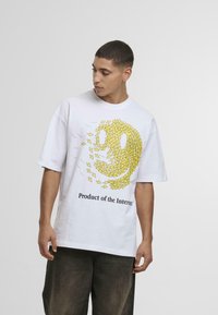 Hvid kortærmet T-shirt med et stort gult smiley ansigt design, der har pile og teksten "Produkt af internettet" nedenunder.