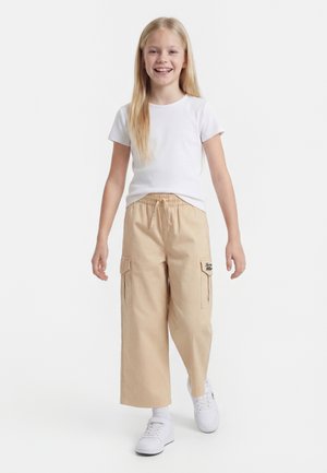 Junges Mädchen lächelt, trägt ein weißes kurzärmeliges Shirt, beige weite Cargohosen, weiße Sneakers, steht vor einem einfarbigen Hintergrund.