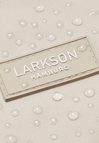 Zbliżenie beżowego materiału z kroplami wody oraz beżową metką "LARKSON HAMBURG" wszytą w powierzchnię.