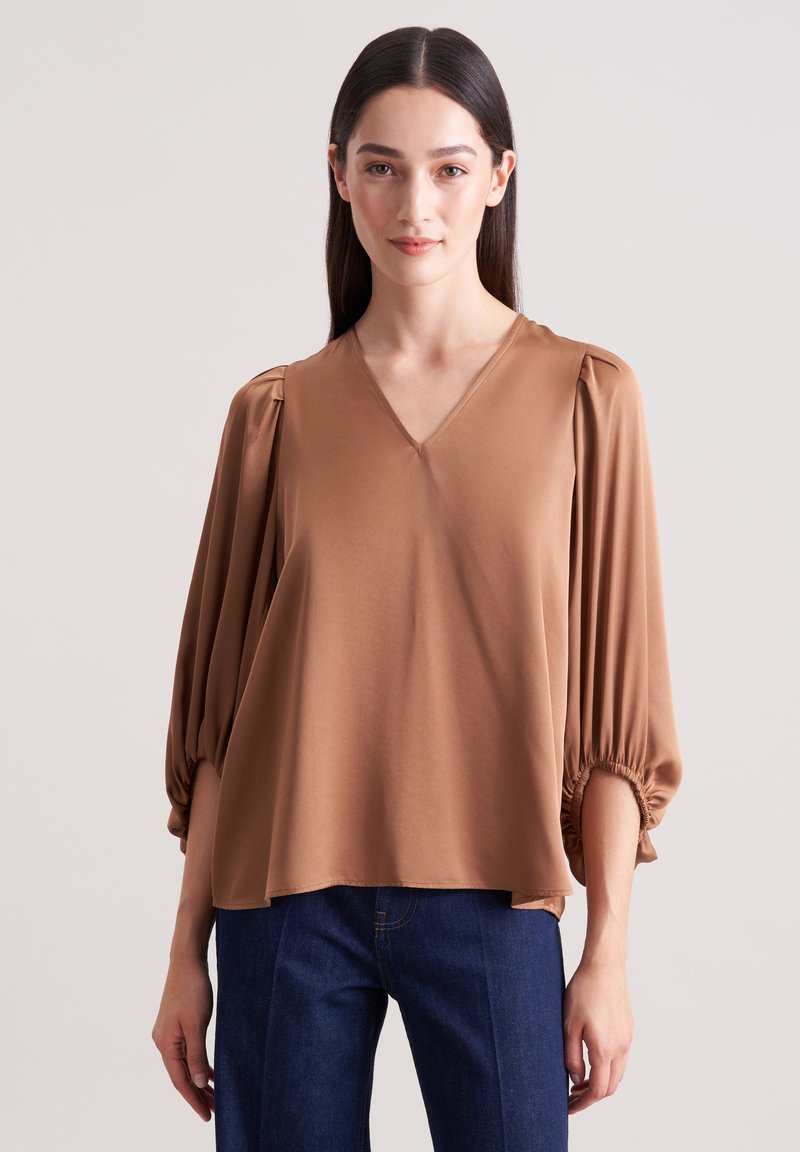 Brun V-hals bluse med puffede ærmer og elastiske manchetter, lavet af glat stof. Sammen med mørkeblå lige ben jeans.