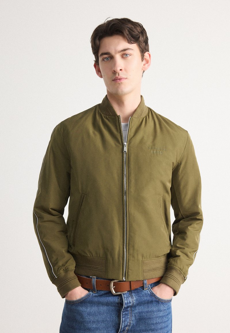 Olive green bomber jacket with zipper closure, elastic cuffs, side pockets, and white stripe accents on sleeves; lightweight fabric.