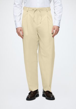 Homme portant un pantalon plissé beige avec une taille à cordon, une chemise blanche boutonnée et des chaussures en cuir marron foncé.