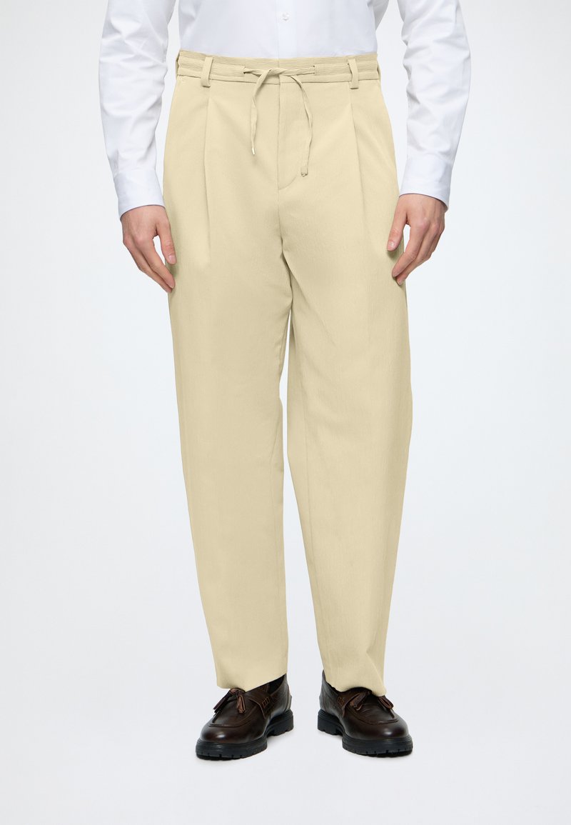 Homme portant un pantalon plissé beige avec une taille à cordon, une chemise blanche boutonnée et des chaussures en cuir marron foncé.