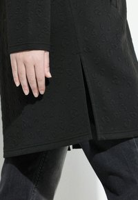 Abrigo negro texturizado con un sutil estampado animal, con una abertura lateral y corte recto, complementado por unos pantalones de mezclilla oscuros.