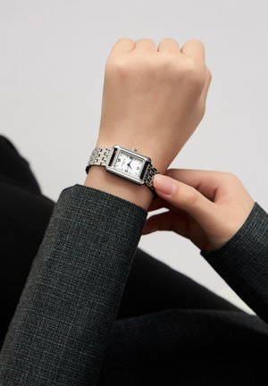 Montre en métal argenté avec un cadran rectangulaire, un cadran blanc, des aiguilles bleues et des chiffres romains. Attachée à un bracelet en métal à maillons.