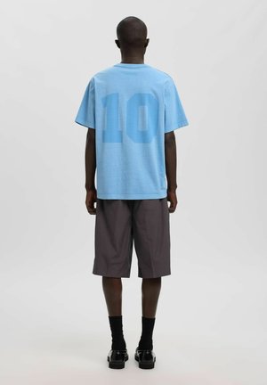 Homme debout, de dos, portant un t-shirt bleu clair avec le numéro 10 dans le dos, un short foncé aux genoux, des chaussettes noires et des chaussures noires.