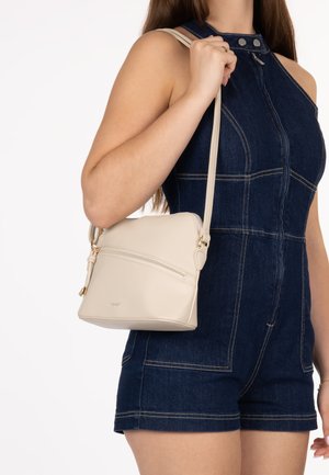 Frau trägt einen dunkelblauen Denim-Overall und hält eine kleine beigefarbene Leder-Schultertasche mit vorderer Reißverschlusstasche.