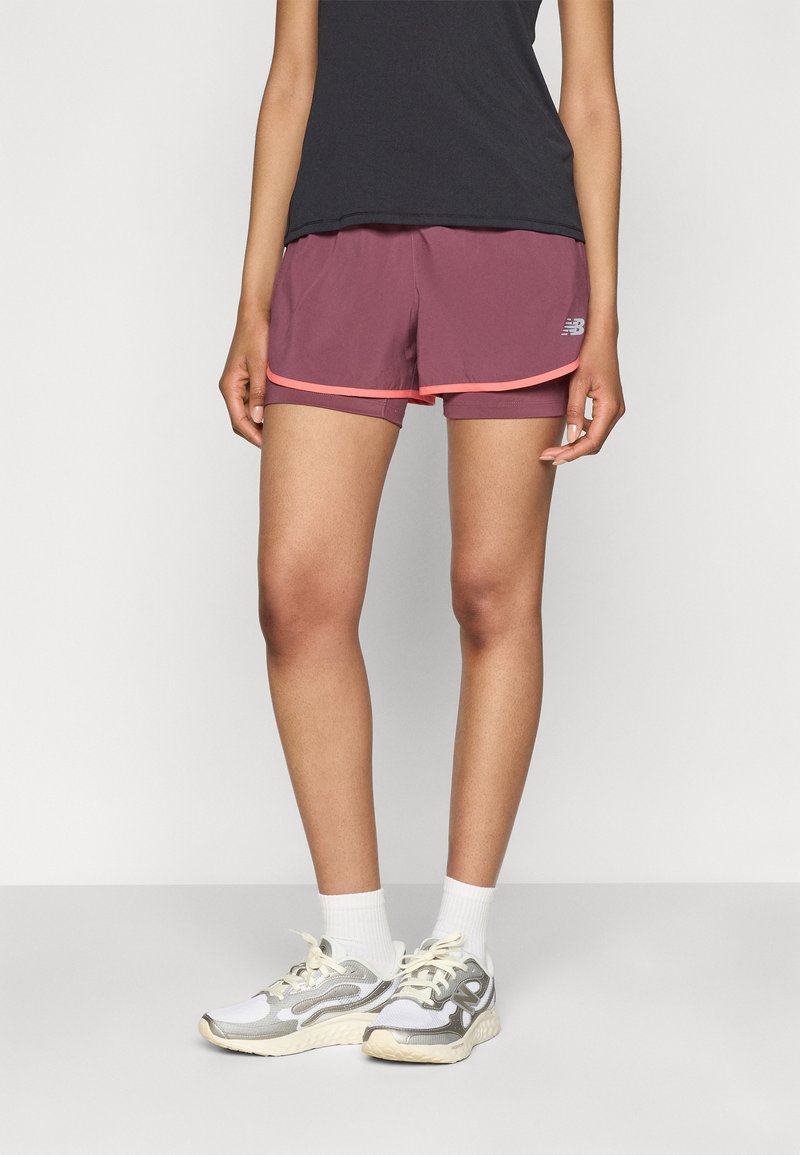 Shorts de sport bordeaux avec une bordure rose, dotés d'une doublure intégrée et d'un logo. Portés avec des baskets grises et blanches et des chaussettes blanches.
