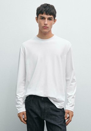 Langärmliges weißes Baumwoll-T-Shirt mit Rundhalsausschnitt, lockerer Passform und leicht abgesenkten Schultern, kombiniert mit dunkelblauen Denim-Jeans.