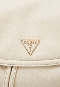 Bolso de cuero sintético color crema con superficie texturizada, con una placa de logo triangular dorada con la inscripción "GUESS USA" en relieve.