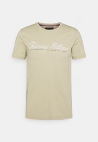 Beżowy bawełniany t-shirt z krótkimi rękawami, okrągłym dekoltem i białym logo „Tommy Hilfiger EST 1985” na przodzie.