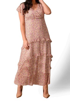 Kobieta ubrana w różową, kwiatową, wielowarstwową suknię maxi z krótkimi rękawami i cielistymi szpiczastymi szpilkami, stojąca z jedną ręką przy twarzy.