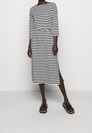 Kobieta ubrana w midi sukienkę w czarno-białe poziome paski, z rękawami do łokcia, pasem w tym samym wzorze, rozcięciem z boku i czarnymi sandałami na platformie.