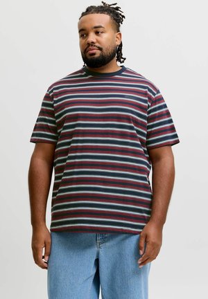 Mand med fletninger iført en t-shirt med vinrøde, hvide og marineblå striber og lyseblå jeans, stående mod en ensfarvet baggrund.