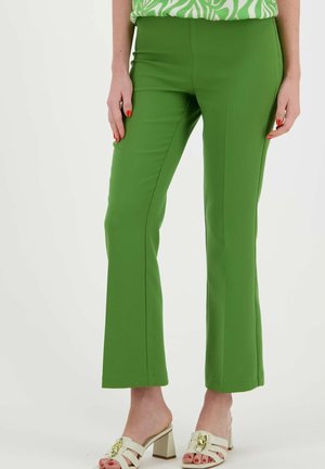 Pantalons tailleur verts en matière extensible, avec une coupe évasée et une texture lisse. Portés avec des sandales à talons blocs de couleur claire.