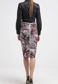 Une femme portant une chemise noire à manches longues et une jupe longueur genou avec des motifs de feuilles blancs et bordeaux, s'éloignant sur un fond blanc uni.