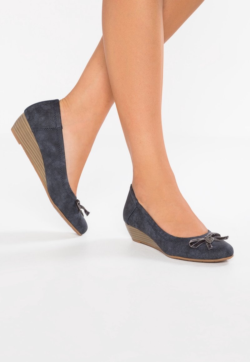 Jambes portant des chaussures compensées en suède gris foncé avec un petit nœud décoratif à l'avant, présentées sur un fond blanc.
