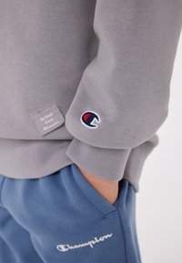 Felpa grigia con polsini a costine, caratterizzata da un piccolo etichetta testuale e un patch con logo in rosso, bianco e blu. Abbinata a pantaloni della tuta blu.