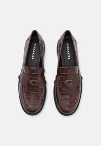 Mocassins en cuir marron avec une texture crocodile, dotés d'une bride rembourrée et d'un logo circulaire à l'avant. Semelles en caoutchouc noir.