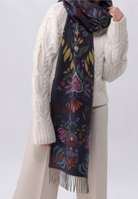 Personne portant un pull crème en maille torsadée tenant une longue écharpe bleu marine avec des motifs floraux colorés et des bords à franges.