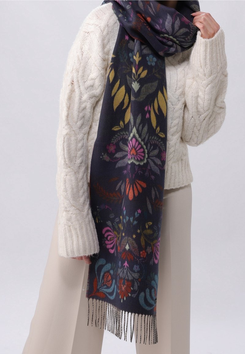 Personne portant un pull crème en maille torsadée tenant une longue écharpe bleu marine avec des motifs floraux colorés et des bords à franges.