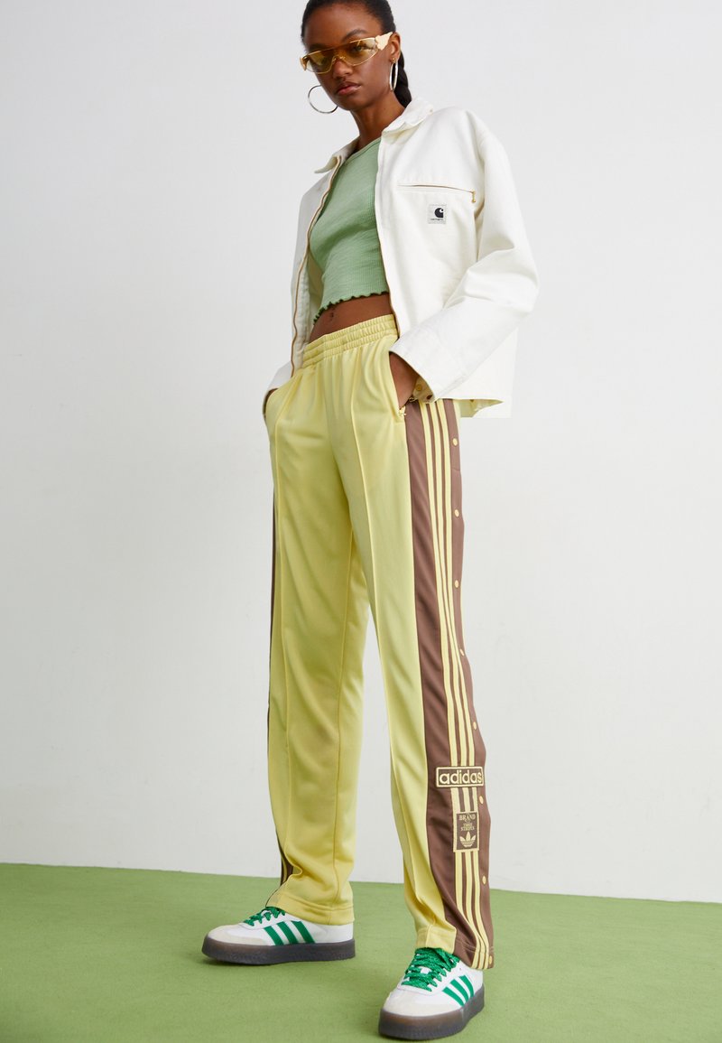 Żółto-brązowe spodnie dresowe z napami, trzy paski po bokach oraz logo Adidas. W zestawie z jasnorozowym topem i białą kurtką.