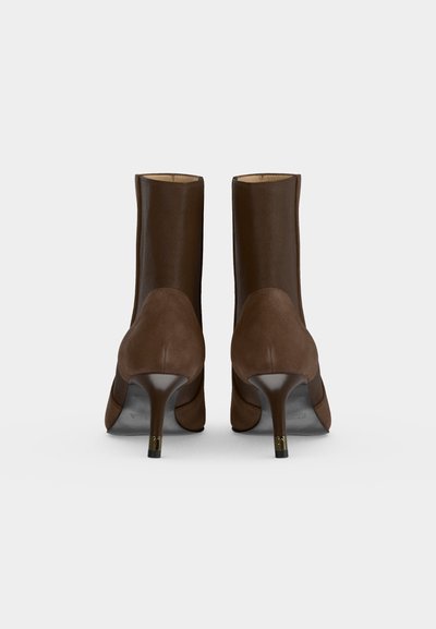 Paire de bottines marron à talons hauts avec dos en daim et talons aiguilles, vues de dos sur un fond clair uni.