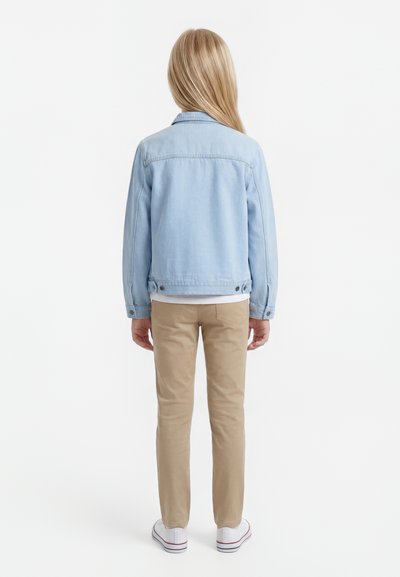 Barn med langt blond hår iført en lyseblå denimjakke, beige bukser og hvide sneakers, der står med fronten væk mod en hvid baggrund.