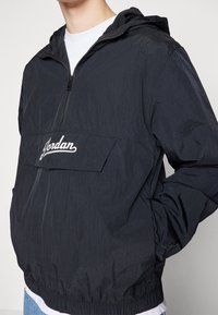 Person som bär en svart huva vindjacka med halvdragkedja och en vit "Jordan"-logotyp broderad på bröstfickans klaff.