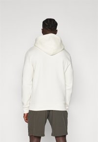 Cremefarbener Hoodie mit großer Kapuze, langen Ärmeln und geripptem Saum. Getragen mit dunkelgrünen Shorts. Glatte Textur und lässige Passform.
