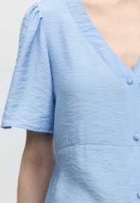 Ανοιχτό μπλε υφασμάτινο μπλουζάκι με κοντά μανίκια με φουσκωτά μανίκια, V-λαιμόκοψη και κουμπιά καλυμμένα με ύφασμα κατά μήκος της μπροστινής πλευράς.