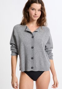 Femme portant un cardigan gris ample à boutons avec un bas noir, se tenant devant un fond blanc uni.